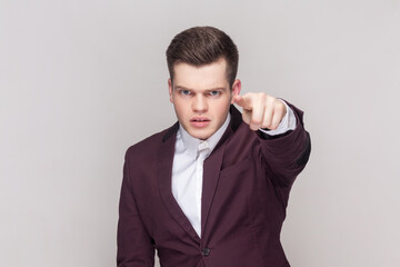 Serious strict handsome handsome young man looks angrily or with judgemental points index finger at camera, wearing violet suit and white shirt. Indoor studio shot isolated on grey background.