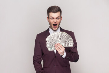 Portrait of surprised excited handsome young man holding big fan of money banknotes, winning lottery, wearing violet suit and white shirt. Indoor studio shot isolated on grey background.