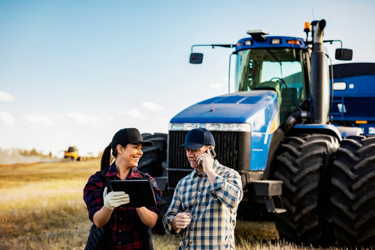A husband and wife using portable wireless devices to manage and monitor their canola harvest and making a call while standing in front of a big tractor; Alcomdale, Alberta, Canada