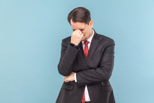 Portrait of sad upset crying man with mustache standing with head down, hearing very bad news, wearing black suit with red tie. Indoor studio shot isolated on light blue background.
