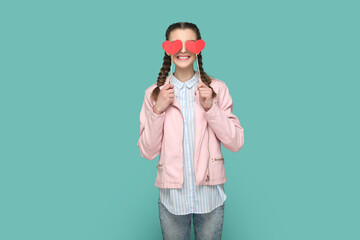 Fototapeta na wymiar Portrait of romantic positive teenager girl with braids wearing pink jacket standing covering her eyes with two red hearts on stick. Indoor studio shot isolated on green background.