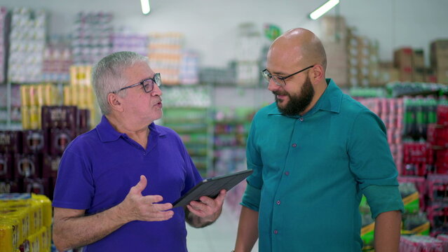 Senior manager of grocery store speaking with staff while looking at tablet device. Two men standing inside local business, job occupation delegation concept
