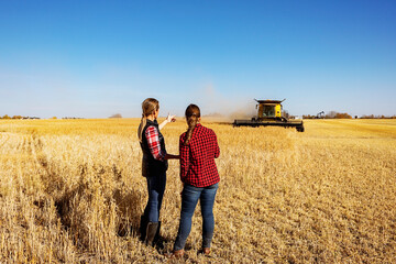 A view from behind of a mature farm woman standing in a field working together with a young woman at harvest time, while a combine is working in the background; Alcomdale, Alberta, Canada