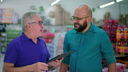 Senior manager of grocery store speaking with staff while looking at tablet device. Two men standing inside local business, job occupation delegation concept