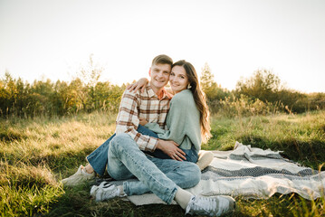 Naklejka na ściany i meble Couple sitting on blanket, hugging in grass in field at sunset. Happy young woman and man, walking spending time together in nature. Concept of family holiday outdoors. Female embrace male on picnic.