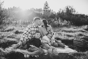 Mother kiss father, daughter, son sitting on blanket, hugging in grass in field at sunset. Children embrace parents. Happy young family walking spending time together in nature. Black and white photo