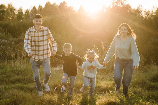 Happy young family with children spending time together, running outside, go in nature at sunset. Mom, dad, daughter and son walk in green grass in spring field. Concept of family holiday outdoors.