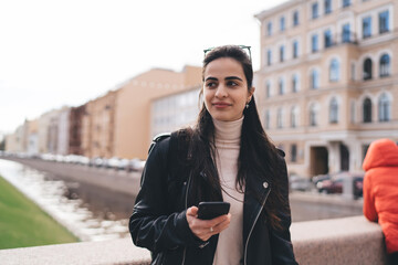 Smiling woman in black jacket standing with smartphone on street