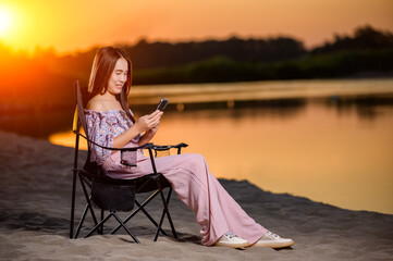 Asian woman sitting on a black chair looking at smartphone on the beach during sunset