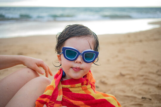 Close-up portrait of young girl wearing swimming goggles warpped in a beach towel and sitting by her mother on the beach; Maui, Hawaii, United States of America