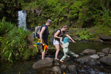 A family carefully walking on a path of volcanic rocks while crossing a stream with a beautiful waterfall in the background on the Road to Hana; Maui, Hawaii, United States of America