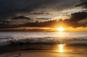 Fototapeta na wymiar Ocean waves along the shoreline of Kamaole 2 Beach at twilight with a golden sun glowing under a cloudy sky and reflecting on the shore; Kihei, Maui, Hawaii, United States of America