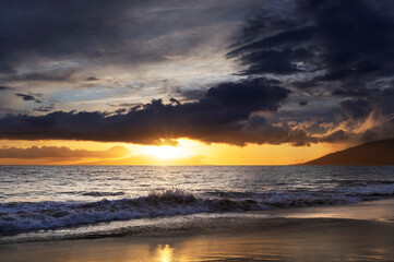 Naklejka na ściany i meble Ocean waves along the shoreline of Kamaole 2 Beach at twilight with a golden sun glowing under a cloudy sky; Kihei, Maui, Hawaii, United States of America
