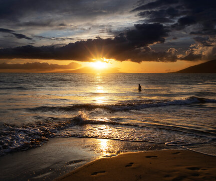 Silhouette of person standing in the water along the shoreline of Kamaole 2 Beach at twilight, watching the golden sun glowing under a cloudy sky; Kihei, Maui, Hawaii, United States of America