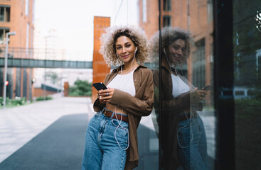 Fototapeta premium Cheerful smiling young ethnic woman with smartphone standing outside building