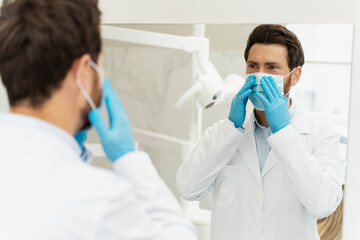 Confident dentist, wearing medical mask and uniform, looking at mirror