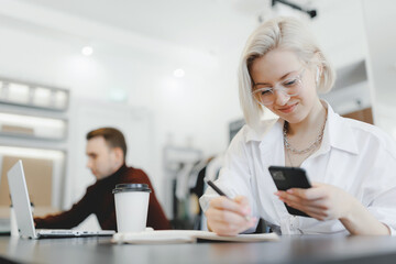 Young female freelancer writes down plan for business in notebook, uses headphones to negotiate with clients