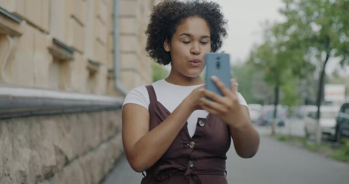 Portrait of cheerful young woman making online video call using smartphone talking and showing city street. Tourism and communication concept.