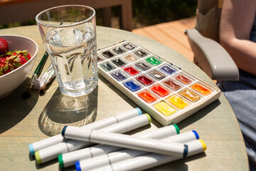 Watercolor granules, paints, watercolor markers, a set of brushes and a glass of water for drawing, on a wooden background, on a sunny day