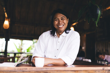 Cheerful Asian man laughing while sitting at table in cafe