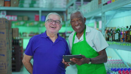 Two senior employees of Grocery store posing for camera smiling inside small business while holding tablet. Older diverse managers of supermarket