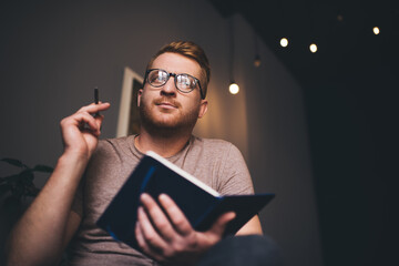 Man in glasses looking away thoughtfully while writing notes in notebook
