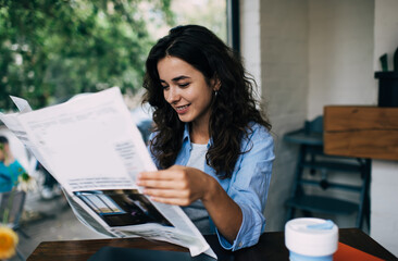 Cheerful Caucasian female with smile on face spending time in street cafe reading news from printed mass media on leisure, happy good looking woman in casual wear holding newspaper checking info