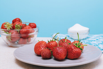 Heap of freshly harvested strawberries on a light table . Strawberries served in a ceramic plate and glass bowl with bowl of sugar.