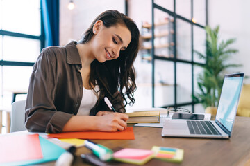 Smiling female student writing on colored paper while studying on laptop