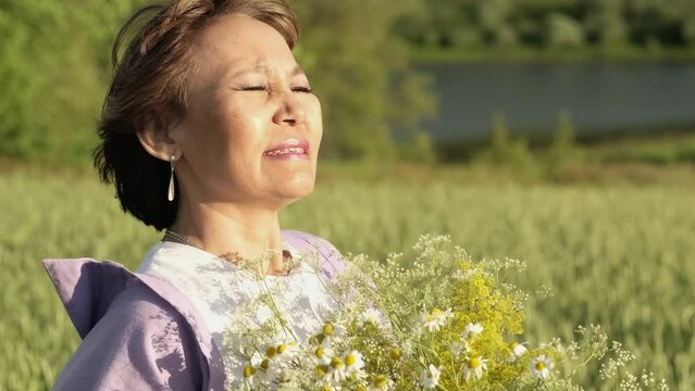 elderly but still attractive Asian woman, breathing in the scent of a wild flower in a bright sunny park. The image conveys happiness and relaxation. Nature as a source of youth