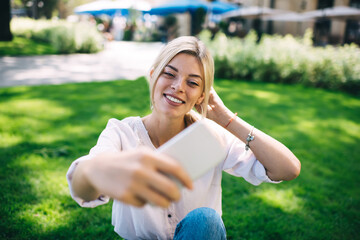 Cheerful female vlogger spending leisure in park for shooting video content for sharing in social media, happy hipster girl posing while clicking selfie photo images on cellular technology