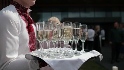 Waiter is holding tray with champagne glasses in her hand. The waiter serves people at the restaurant. Alcohol drinks in wine glasses. Night party, holidays event outdoor. People drinking.