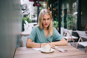 Portrait of young intelligent student with caffeine beverage smartphone and education textbook for learning looking at camera during free time in street coffee shop, smart woman with notepad posing