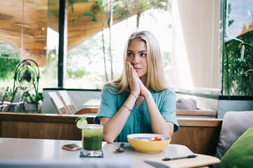 Contemplative hipster girl with matcha beverage and plate of organic fruit muesli thoughtful looking away and thinking about portion calorie in cafe interior, pondering woman have healthy breakfast