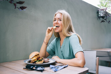 Young Caucasian female having fun while eating appetizing burger with beef patty sitting at table...