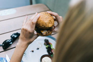 Female customer eating tasty American burger with sesame bread and vegan meat, unrecognizable woman enjoying leisure time for have lunch with delicious caloric hamburger during cheat meal on diet