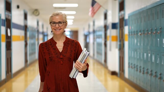Closeup portrait of pretty mature woman teacher holding books walking down an empty school hallway.