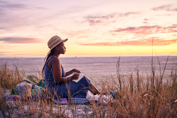 Black woman, relax and book on beach in sunset with diary or journal for vacation in nature outdoors. African female person relaxing on ocean coast with notebook in morning sunrise and holiday travel