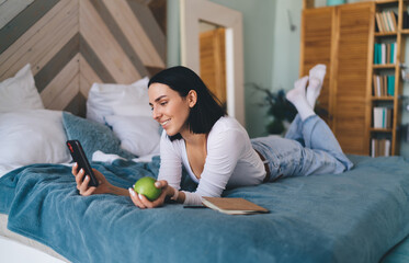 Smiling woman using smartphone and resting on bed