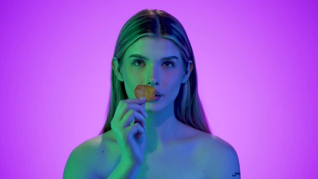 Blondynka na fioletowym tle pokazuje chrupkę chipsa i wkłada ją do ust. Twarz oświetlona jest zielonym i niebieskim światłem z dw&oacute;ch stron. Ujęcie kolorowe, nowoczesne, z nutą stylizacji