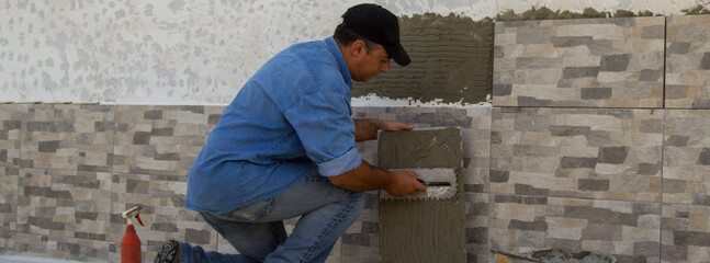Image of a handyman tiler who with a spatula spreads the glue for laying the tiles on a house wall. Do-it-yourself work and home renovation. Horizontal banner 
