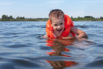 Little, happy boy in orange life vest swimming in lake waves. (selective DOF)