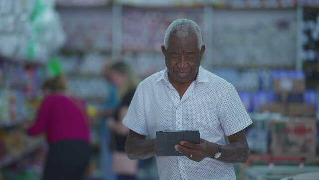 Confused senior man using tablet device inside small business store. One older Black man scratching forehead while staring at screen, engaged with modern technology
