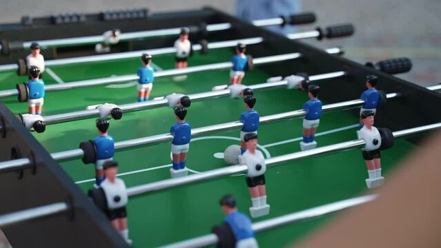 Teenager puts ball on green kicker field to start game and begins to rotate handles with fixed players, close-up. Playing table football in open air and entertainment for holiday guests.