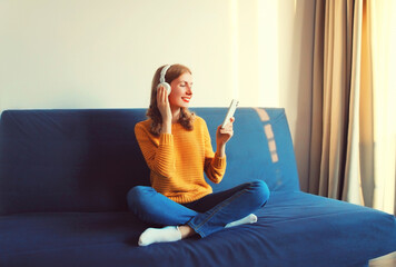 Happy smiling young woman listening to music with wireless headphones looking at smartphone sitting on the couch at home