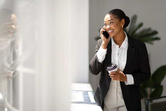 Happy black woman in suit having phone conversation and drinking coffee, female boss standing in office near window