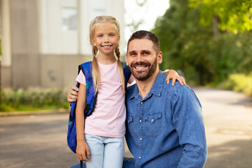 Obraz premium Back to school concept. Little girl with school bag embracing father near school, smiling together at camera
