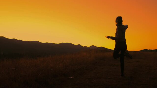 Girl dancing against bright orange sunset sky. Slim young girl silhouette having fun, moving, raise hands up, listening music in mountain field. Back view. Slow motion 4K