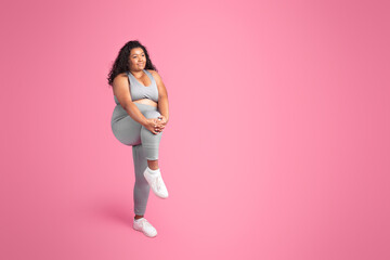 African american chubby lady in sportswear doing leg warm up before training on pink background,...