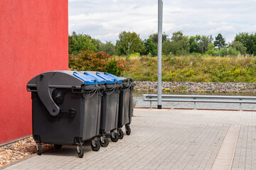 Black plastic trash cans closed on a chain stand near a large red building.
